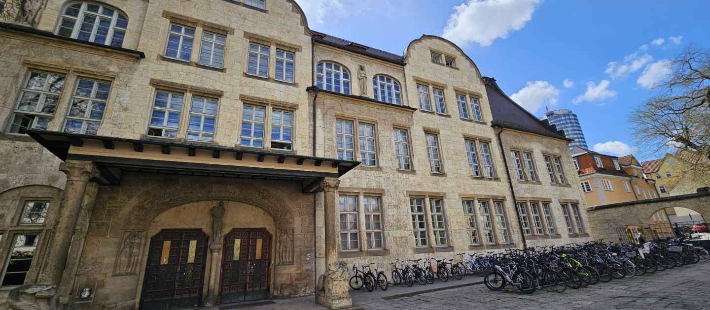 Historisches Sandsteingebäude mit vielen Fenstern und zwei Eingängen, davor zahlreiche geparkte Fahrräder unter blauem Himmel