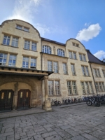 FSU Jena-Eingang-JenaKultur-Christoph-Litwinov Historisches Sandsteingebäude mit vielen Fenstern und zwei Eingängen, davor zahlreiche geparkte Fahrräder unter blauem Himmel
