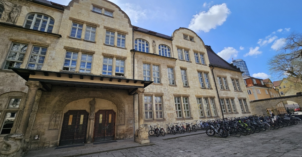 Historisches Sandsteingebäude mit vielen Fenstern und zwei Eingängen, davor zahlreiche geparkte Fahrräder unter blauem Himmel
