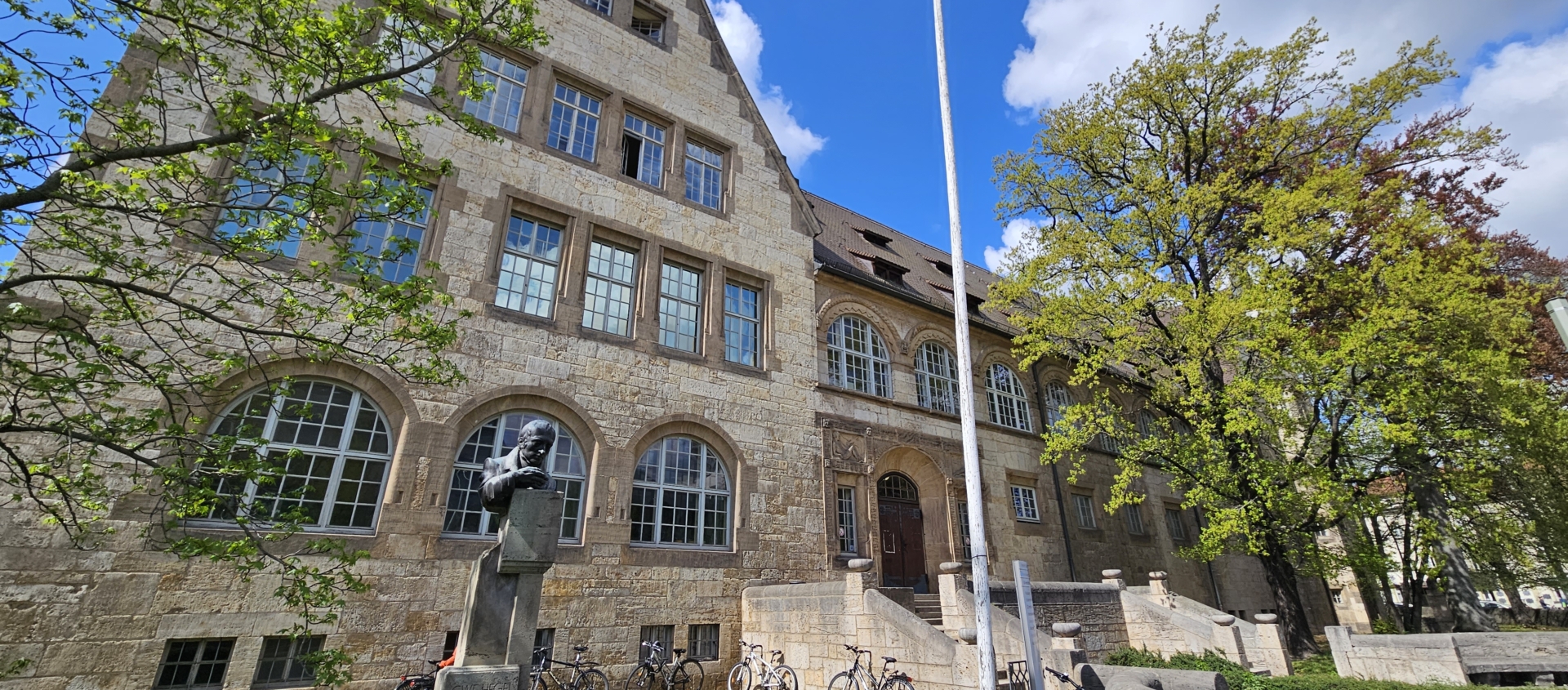 Historisches steinernes Gebäude mit großen Fenstern, Treppe zum Eingang und einer Statue im Vordergrund, umgeben von Bäumen und Fahrrädern