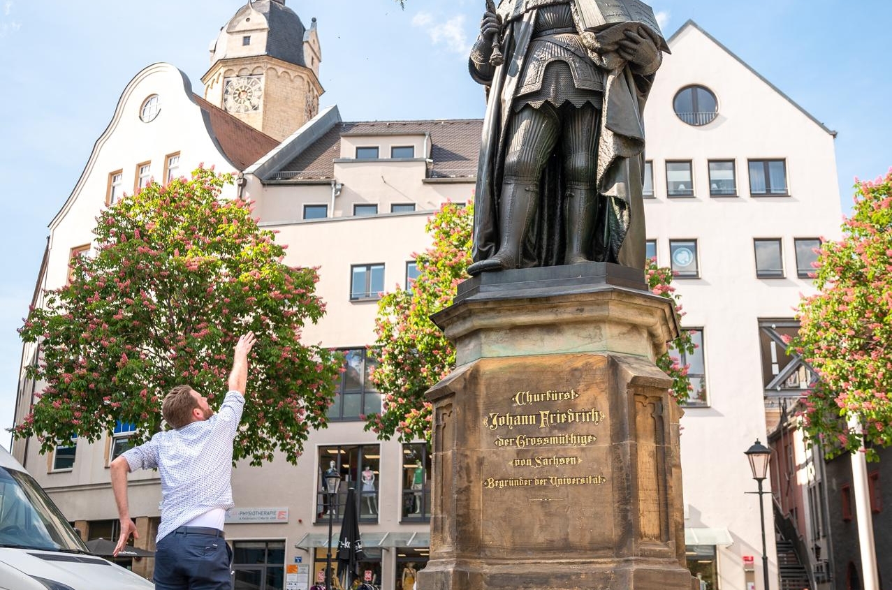 Student wirft Kranz zu Statue von Kurfürst Johann Friedrich auf dem Jenaer Marktplatz bei sonnigem Himmel