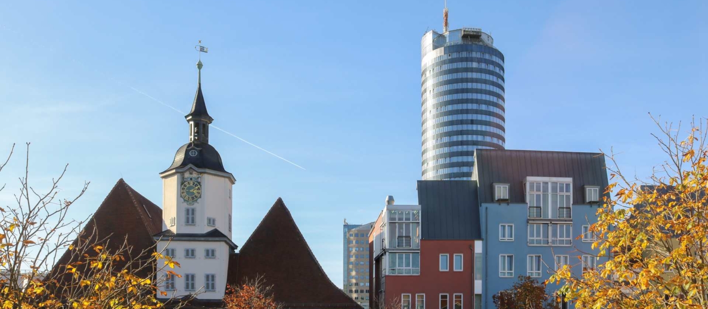 Historisches Rathaus Jena mit weißem Uhrenturm und roten Dächern, umgeben von bunten Häusern und herbstlichen Bäumen, im Hintergrund der Jentower