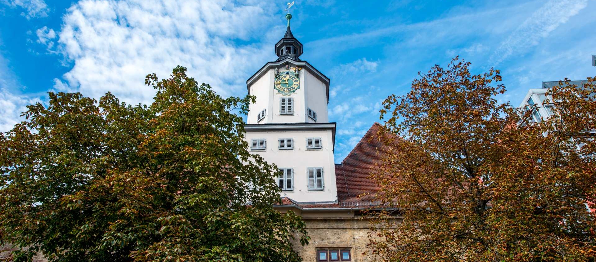 Historisches Rathaus in Jena mit Uhrenturm, umgeben von Bäumen mit herbstlichen Blättern unter blauem Himmel mit Wolken