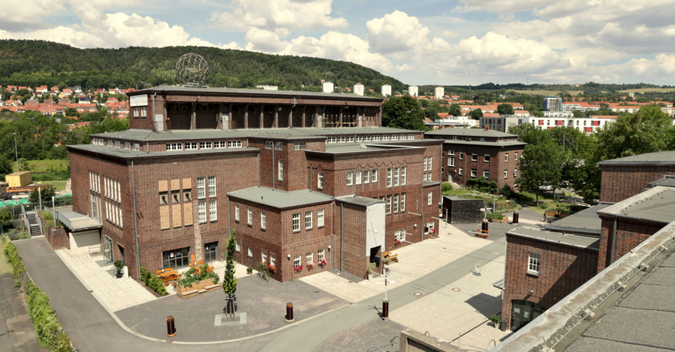 Mehrere mehrstöckige Backsteinbauten auf dem Gelände der imaginata in Jena mit umliegenden Straßen und bewaldeten Hügeln im Hintergrund