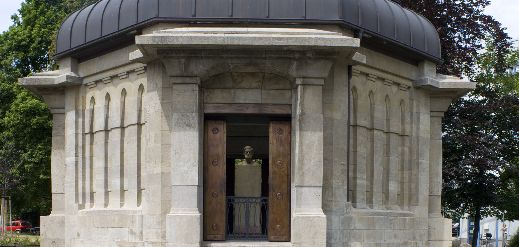 Das Ernst-Abbe-Denkmal zeigt eine steinerne, achteckige Gedenkhalle mit bronzenen Türen und einer Büste im Inneren, umgeben von Bäumen.