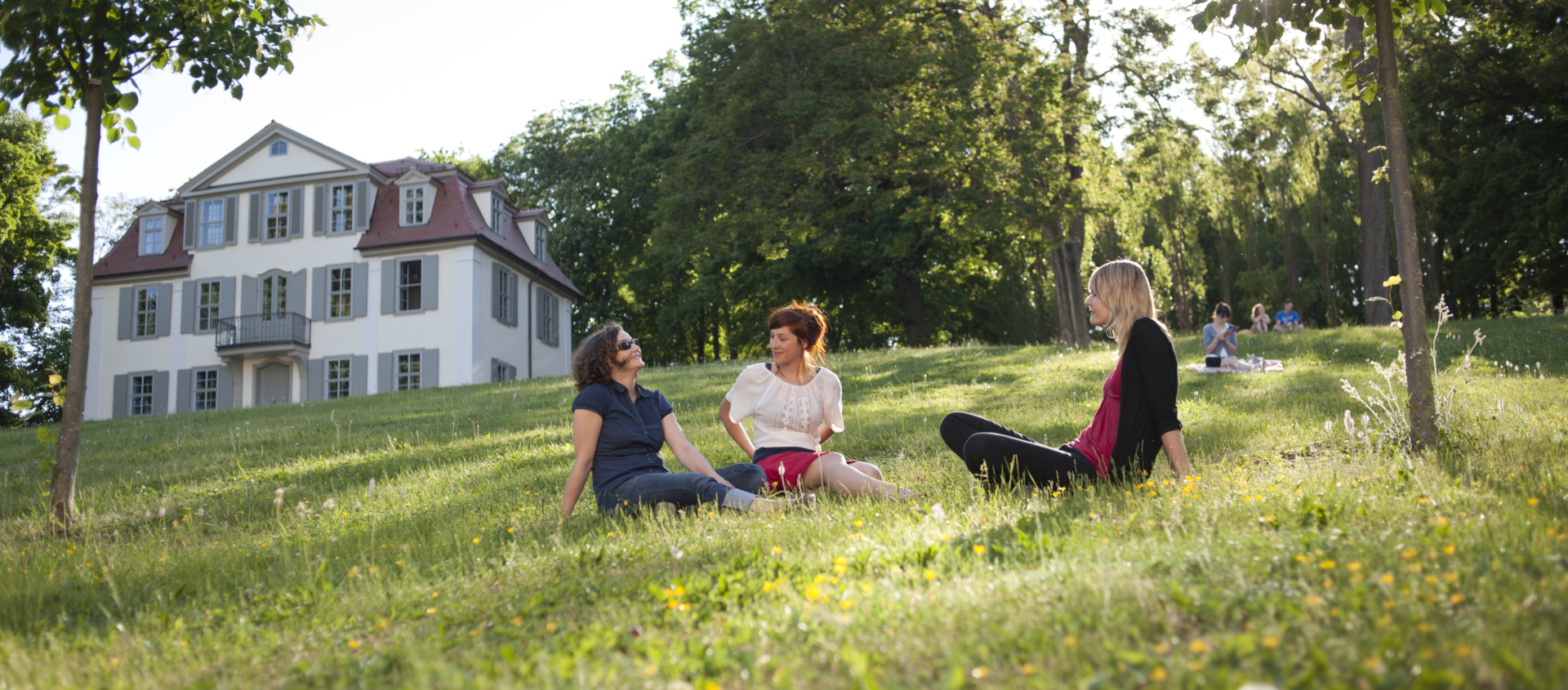 Drei Frauen sitzen auf einer Wiese und unterhalten sich entspannt, im Hintergrund das Griesbachsche Gartenhaus umgeben von Bäumen