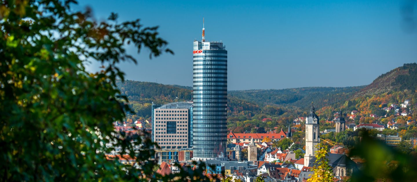 Jentower in Jena als hoher, runder Wolkenkratzer mit umliegenden Gebäuden und bewaldeten Hügeln im Hintergrund