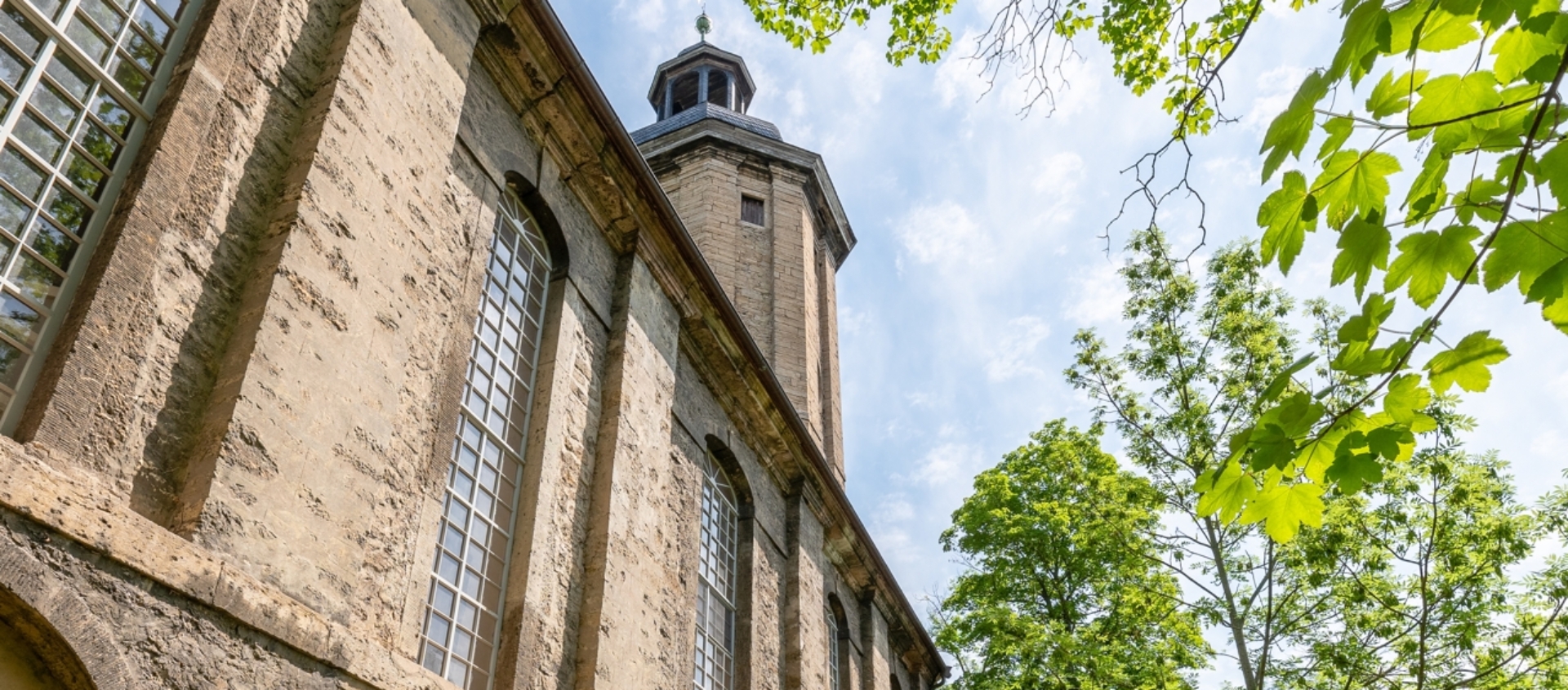 Blick entlang der Außenwand der Friedenskirche in Jena mit hohen Fenstern, Nischen mit Statuen und einem schmalen Weg neben grünen Bäumen und Gras unter blauem Himmel
