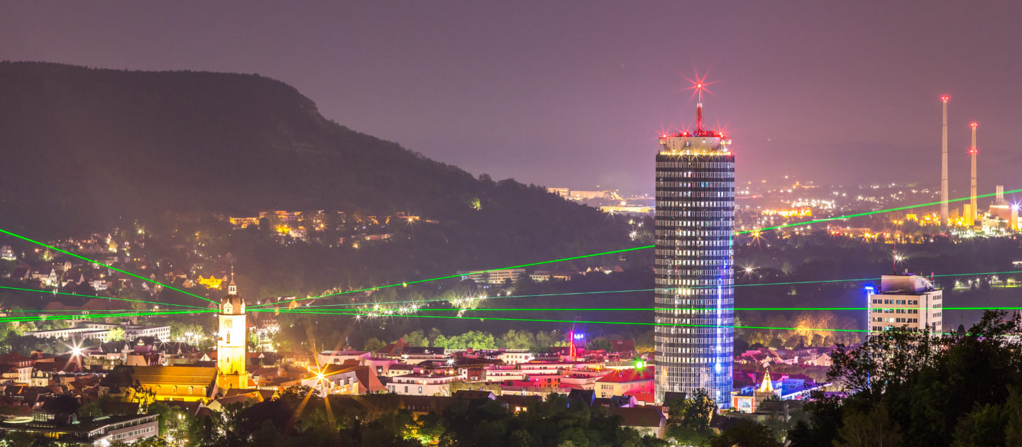 Nächtliche Stadtansicht von Jena mit dem markanten JenTower, umgeben von beleuchteten Gebäuden und grünen Laserstrahlen am Himmel