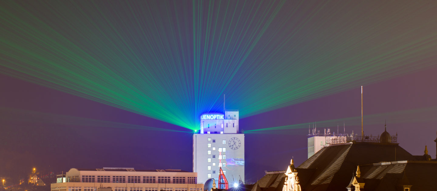 Nachthimmel über Jena mit beleuchtetem Turm der Jenoptik und grünen sowie blauen Laserstrahlen, im Vordergrund historische Gebäude