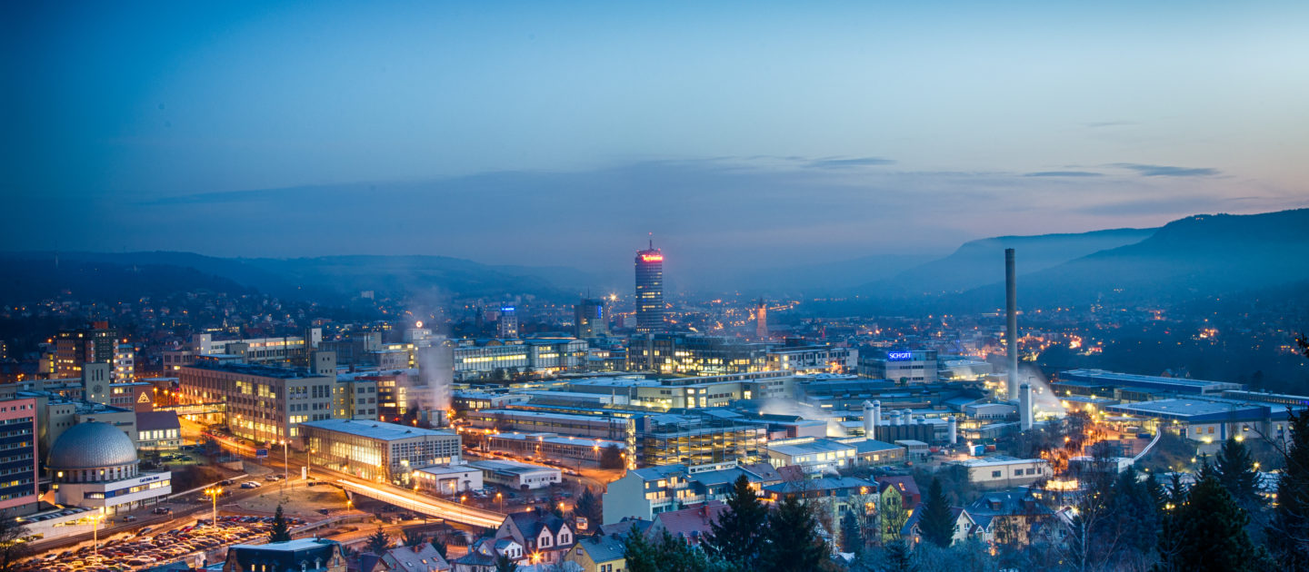 Blick auf die abendliche Stadt Jena mit beleuchteten Gebäuden, Industrieanlagen und Hügeln im Hintergrund
