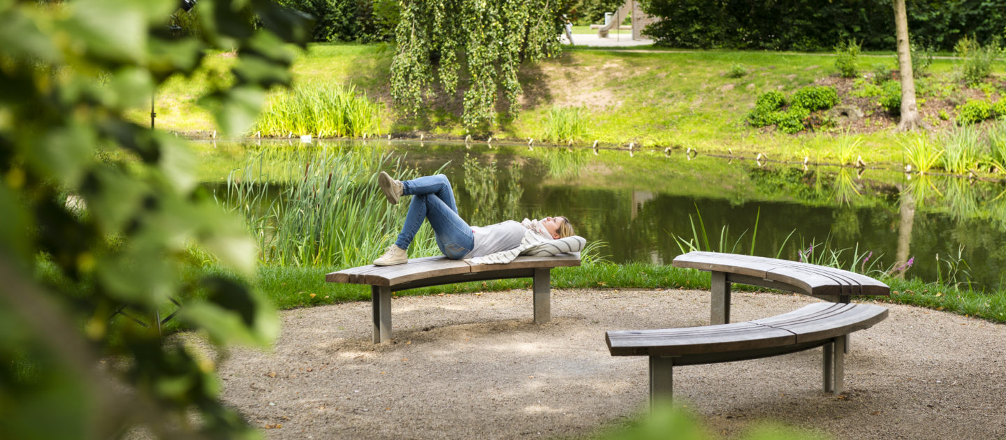 Person liegt entspannt auf einer halbkreisförmigen Holzbank am Teich im Paradiespark in Jena