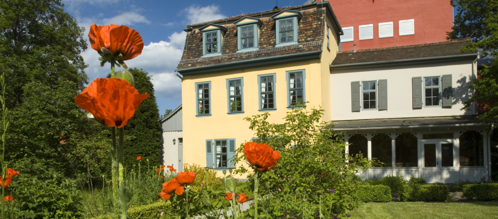 Schillers Gartenhaus in Jena mit gelb gestrichenem Gebäude, blühenden Mohnblumen im Vordergrund und gepflegtem Rasen unter blauem Himmel
