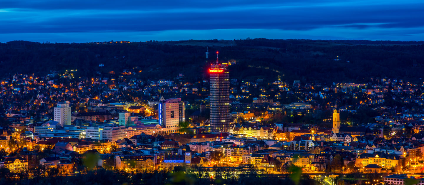 Nächtliche Stadtansicht von Jena mit beleuchtetem JenTower und umliegenden Gebäuden vor bewölktem Himmel