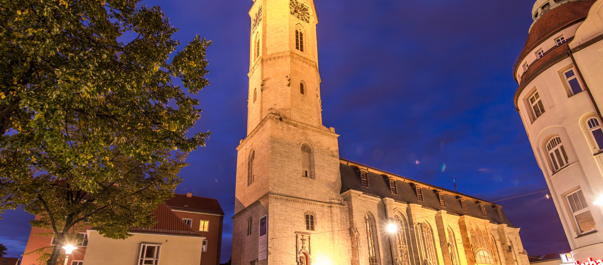 Die beleuchtete Stadtkirche Jena bei Nacht mit hohem Turm, umgeben von Gebäuden und Bäumen unter einem dunklen Himmel