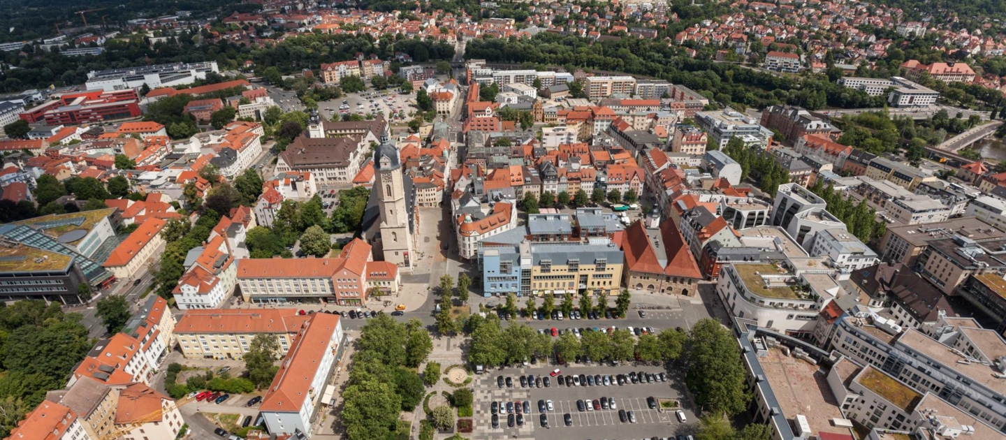 Luftaufnahme von Jena mit Blick auf den Eichplatz, umgeben von historischen Gebäuden, einem großen Parkplatz und bewaldeten Hügeln im Hintergrund