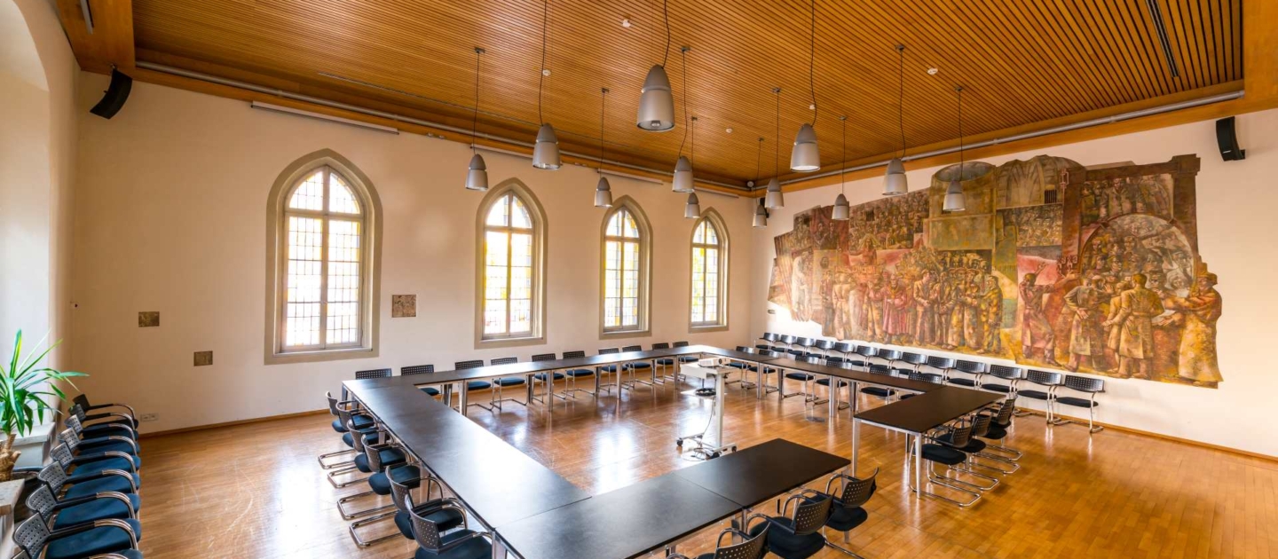 Plenarsaal im historischen Rathaus mit U-förmigen schwarzen Tischen, schwarzen Stühlen, hohen gotischen Fenstern und großer Wandmalerei