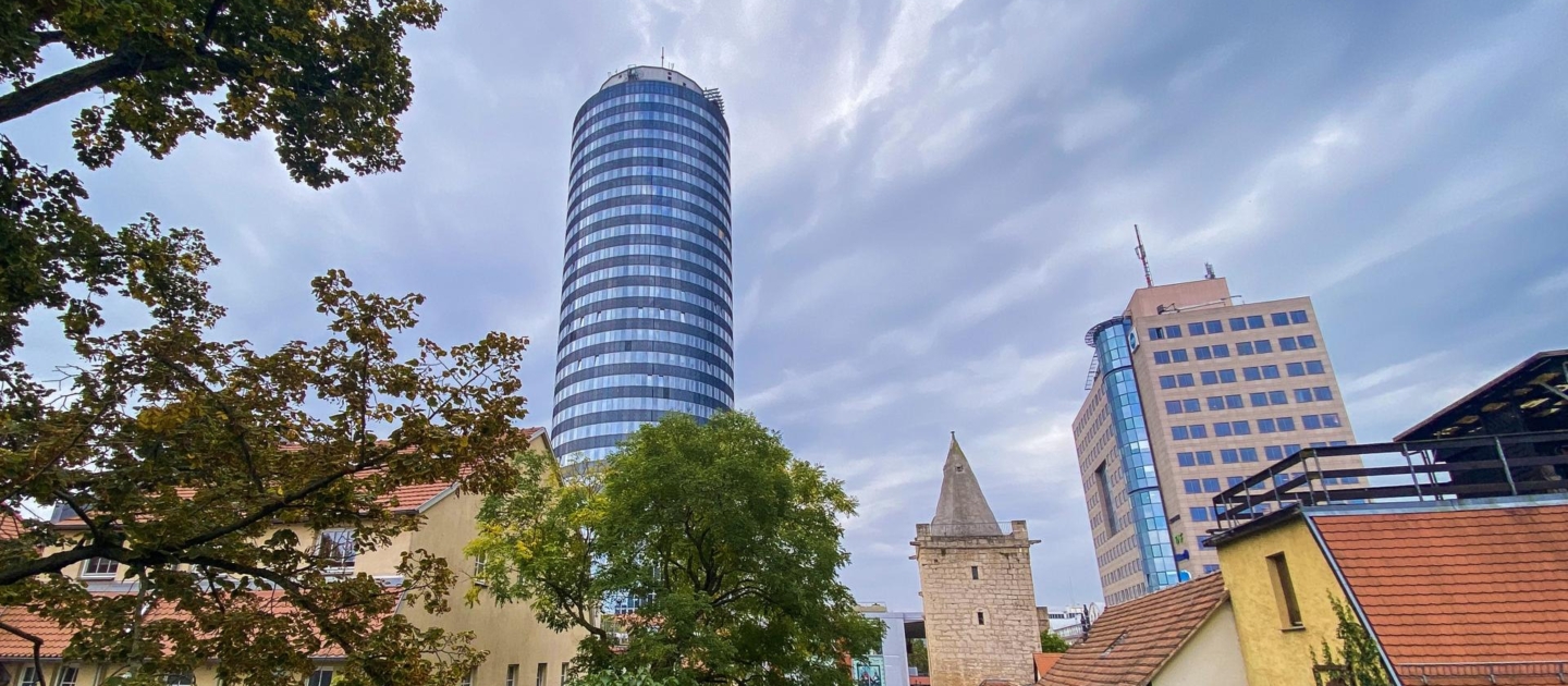 Blick auf den Jentower und das Johannistor in Jena mit Bäumen und historischen sowie modernen Gebäuden unter bewölktem Himmel