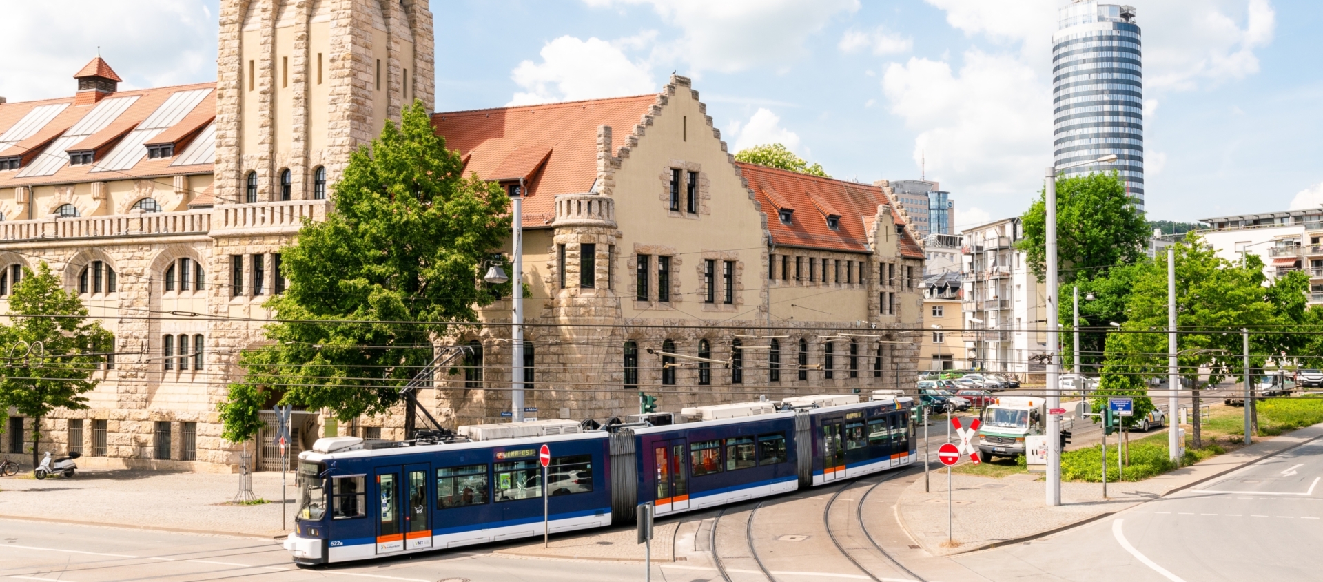 Historisches Volksbad-Gebäude in Jena mit rotem Ziegeldach und Turm, davor fährt eine blaue Straßenbahn auf Schienen