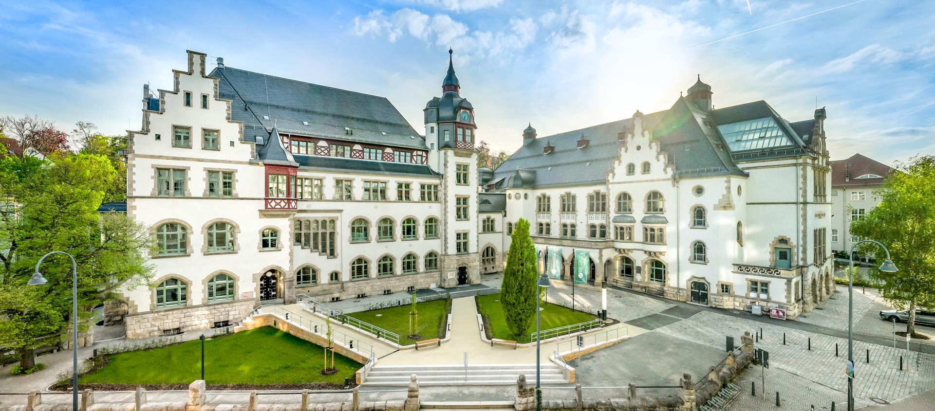 Historisches Volkshaus Jena mit weißer Fassade, Giebeln und Turm, umgeben von Grünflächen und gepflastertem Vorplatz bei sonnigem Himmel