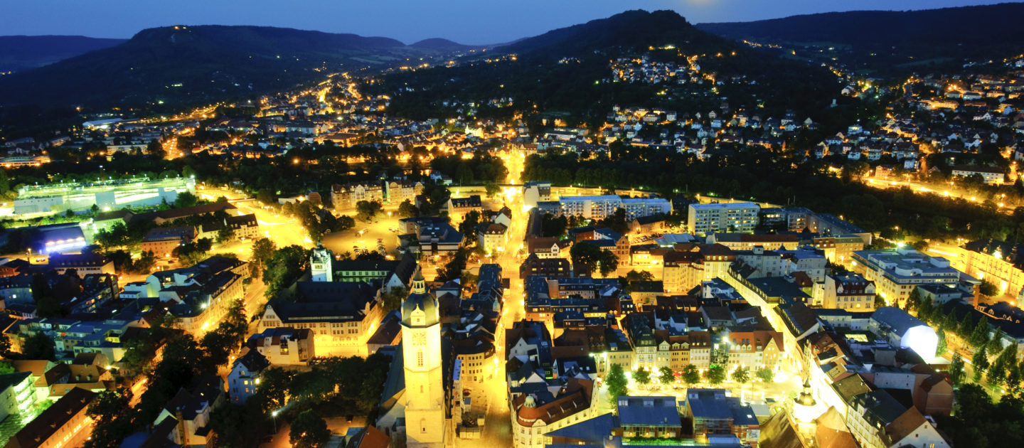 Nachtaufnahme der beleuchteten Innenstadt von Jena mit dem markanten Turm des Rathauses und umliegenden Gebäuden vor bewaldeten Hügeln und einem hellen Mond am Himmel