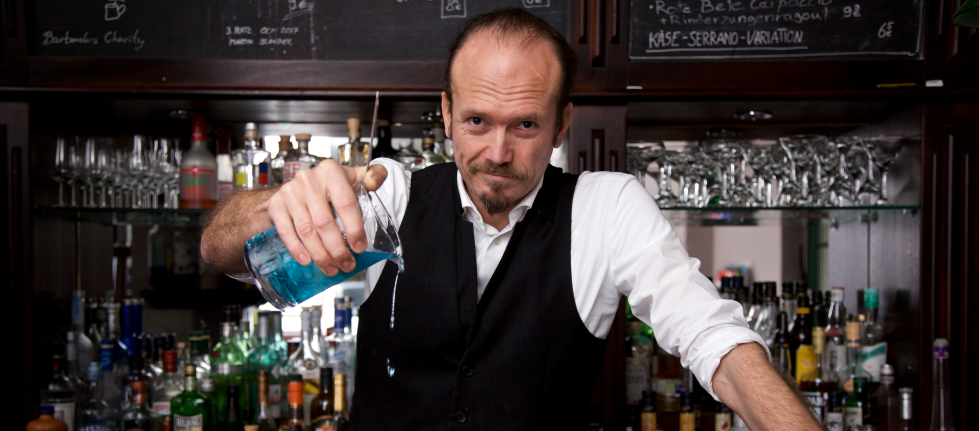 Danny Müller, Chef der Weintanne Jena, gießt blauen Cocktail aus einem Messbecher in zwei Cocktailschalen an einer Bar mit zahlreichen Spirituosenflaschen im Hintergrund.