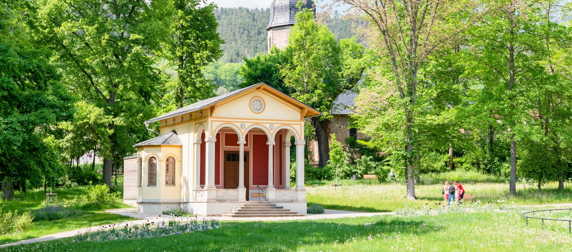 Teehäuschen im Drackendorfer Park in Jena, umgeben von Bäumen, mit der Kirche im Hintergrund und Spaziergängern auf einem Weg
