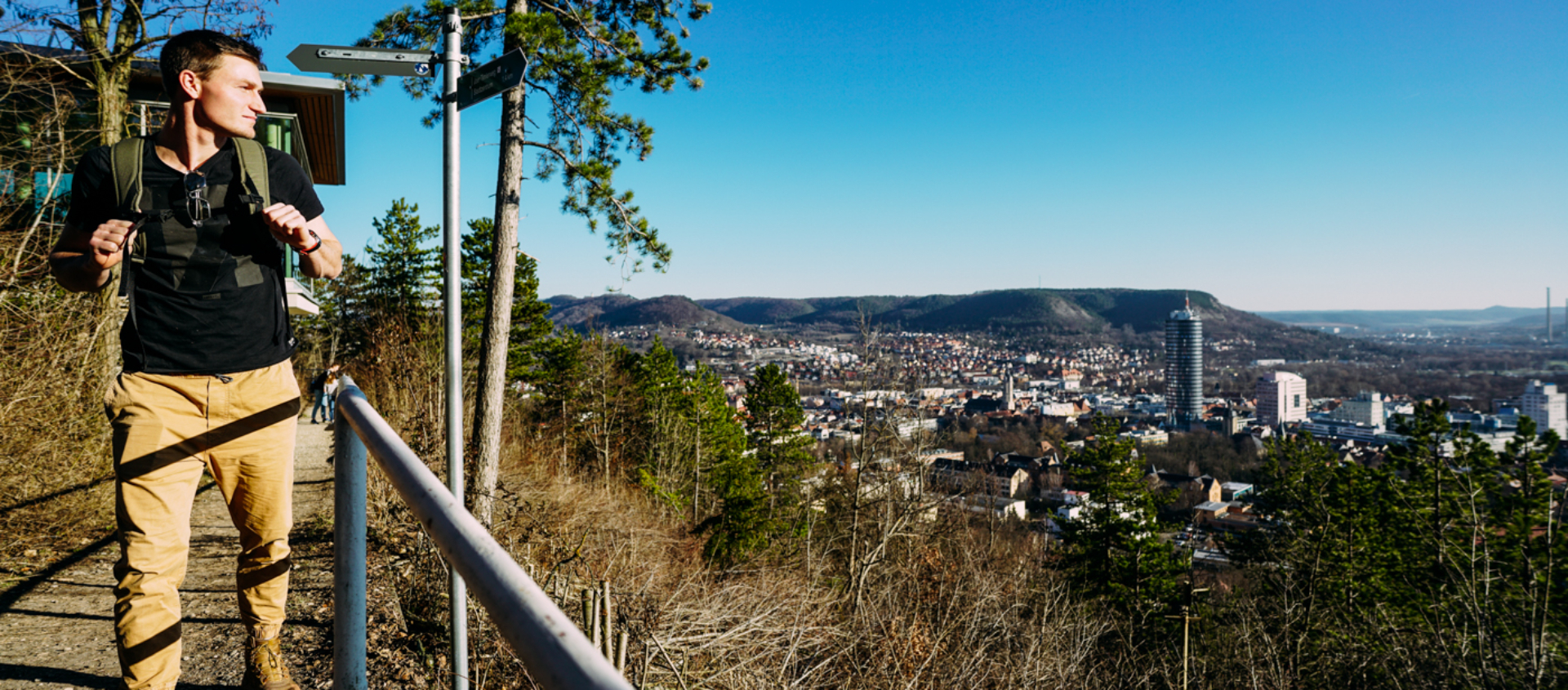 Wanderer mit Rucksack auf einem Hügel mit Blick auf die Stadt Jena und die Saale im Hintergrund