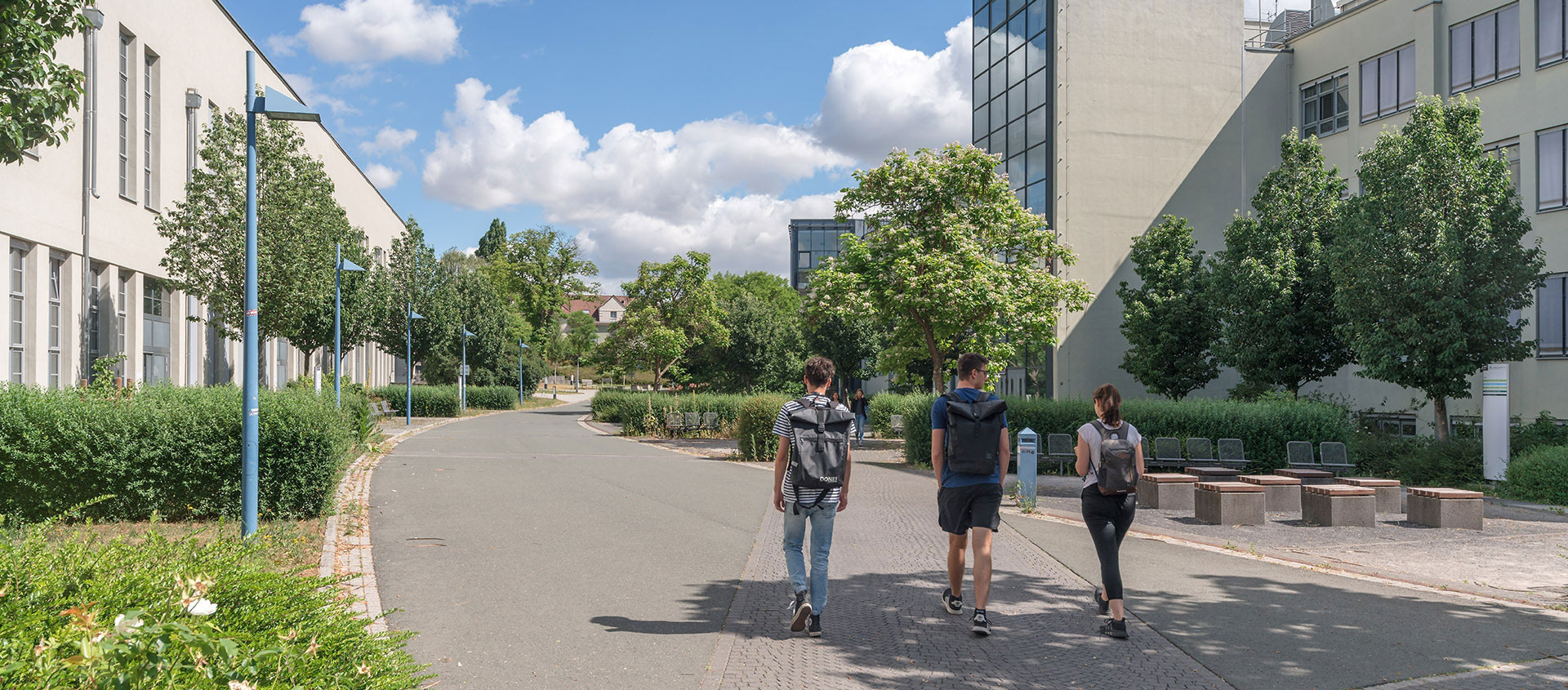 Drei Studierende gehen auf dem Campus der Ernst-Abbe-Hochschule zwischen modernen Hochschulgebäuden bei sonnigem Wetter