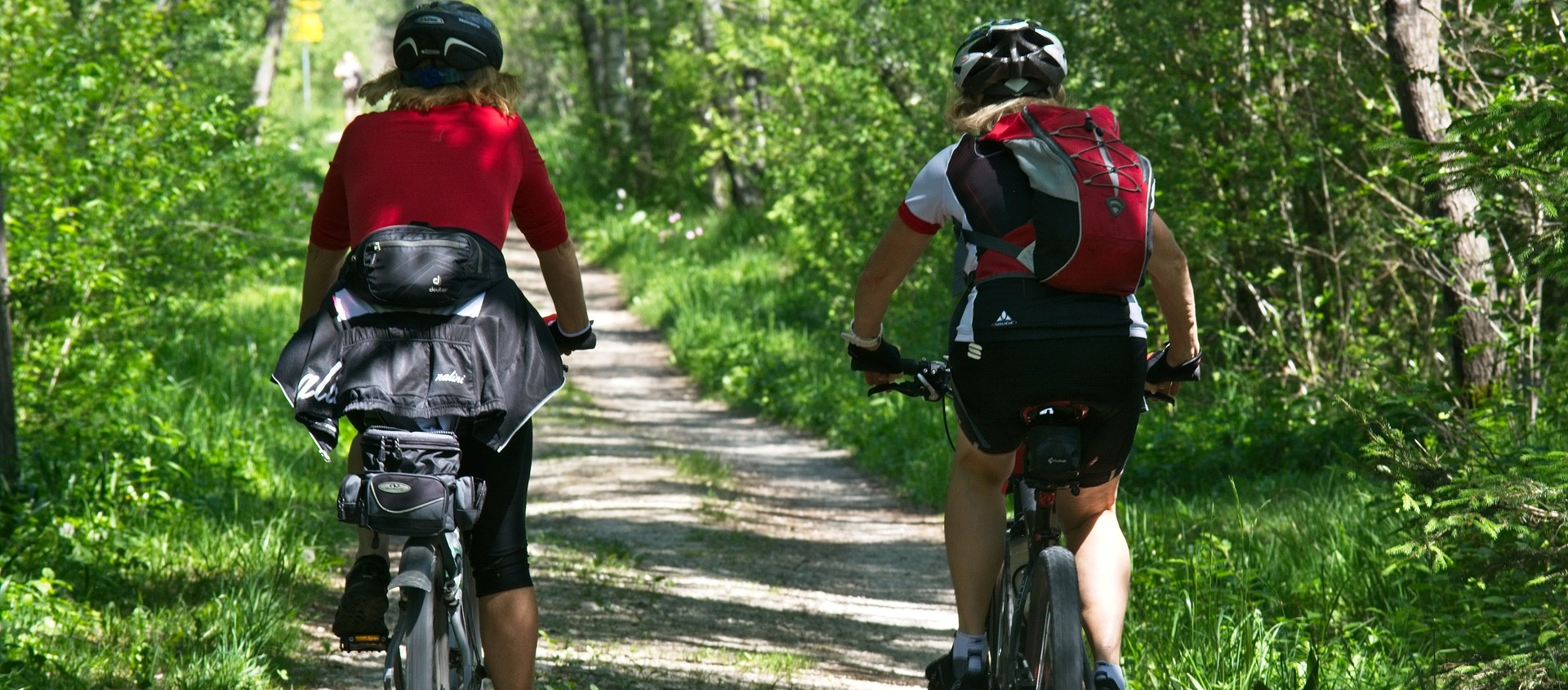 Zwei Radfahrer mit Helmen und Rucksäcken fahren auf einem schmalen Waldweg durch grünes Laub