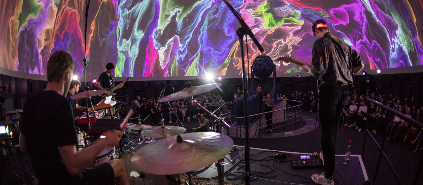 Musiker spielen Schlagzeug und Gitarre auf Bühne im Planetarium mit bunten, abstrakten Projektionen an der Kuppel während des Fulldome Festivals