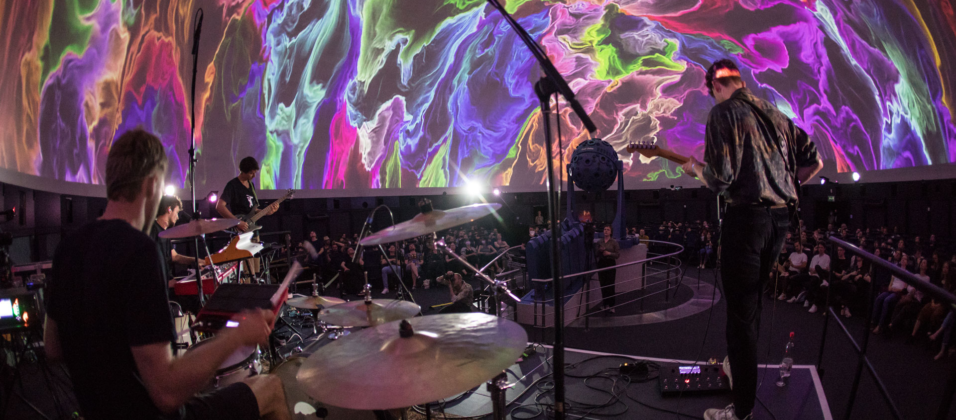 Musiker spielen Schlagzeug und Gitarre auf Bühne im Planetarium mit bunten, abstrakten Projektionen an der Kuppel während des Fulldome Festivals