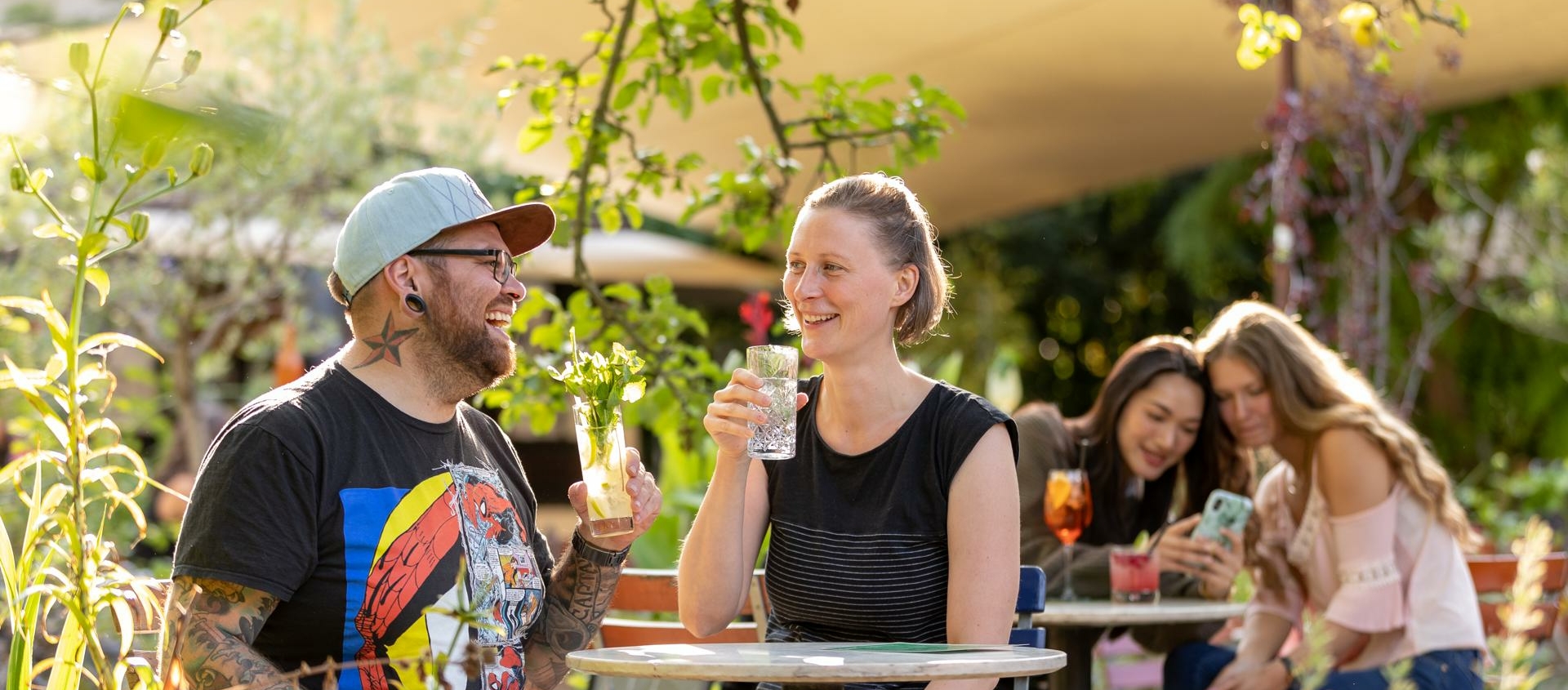 Zwei Personen sitzen an einem runden Gartentisch mit Getränken, im Hintergrund sitzen zwei weitere Personen auf einer Bank unter Bäumen