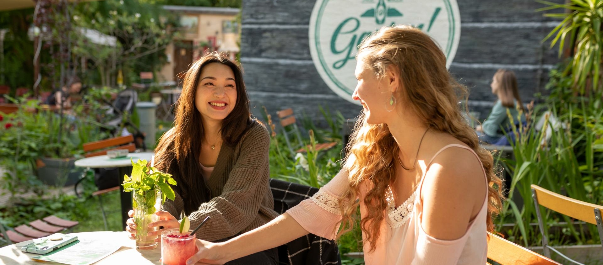 Zwei Frauen sitzen an einem runden Gartentisch mit Getränken, im Hintergrund ein Holzgebäude mit rundem Schild und dem Schriftzug 'Garten'.