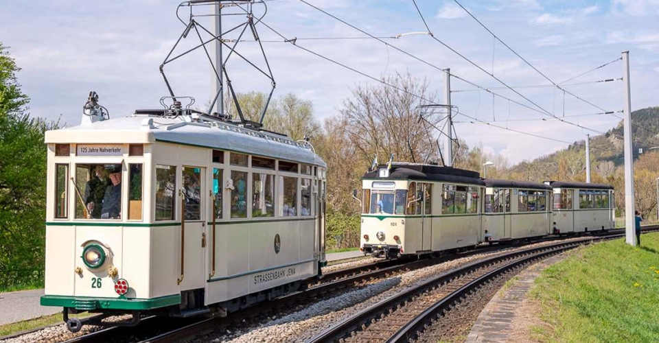 Historischer Triebwagen mit der Nummer 26 und historischer Dreiwagenzug auf einer Gleisstrecke im Freien unter Oberleitungen in Jena