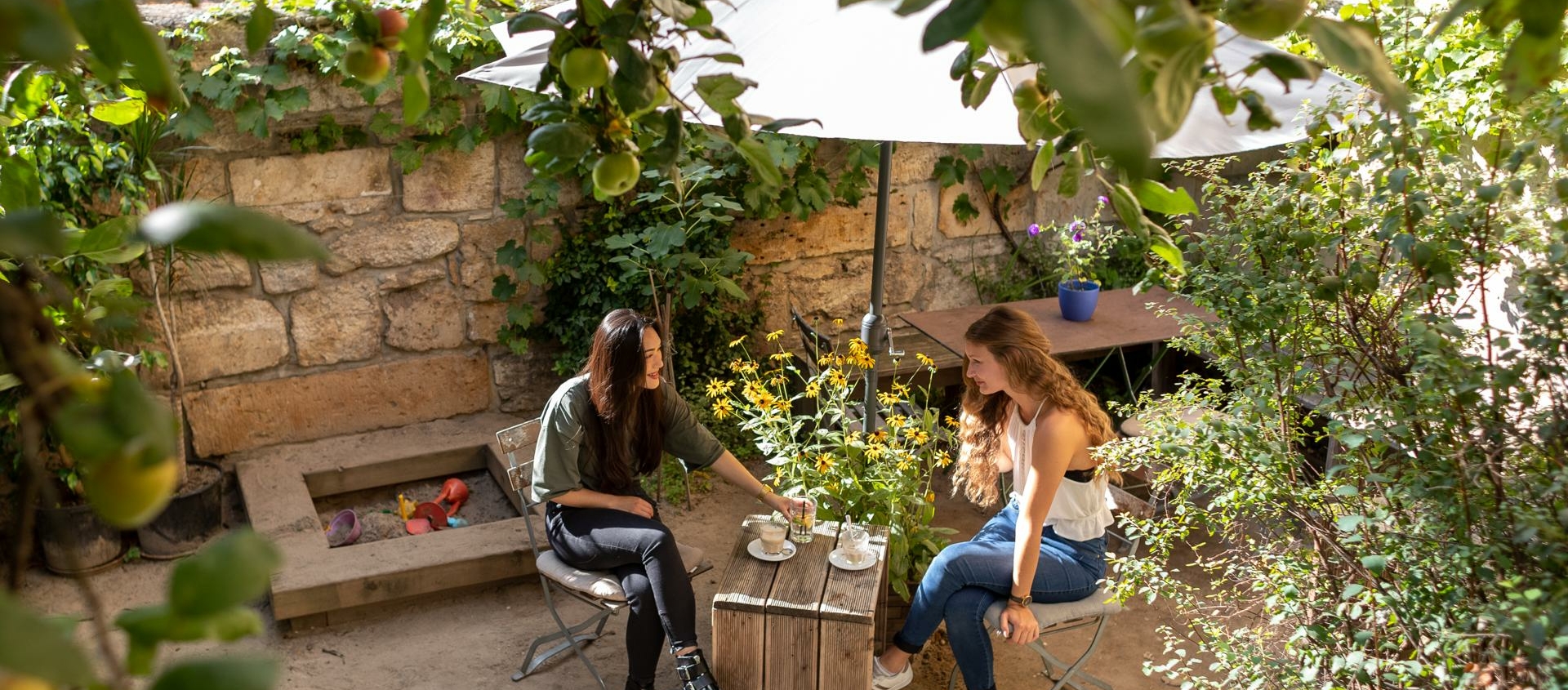 Zwei Frauen sitzen an einem kleinen Holztisch mit Tassen in einem begrünten Innenhof mit Steinmauer und Apfelbaumzweigen im Vordergrund