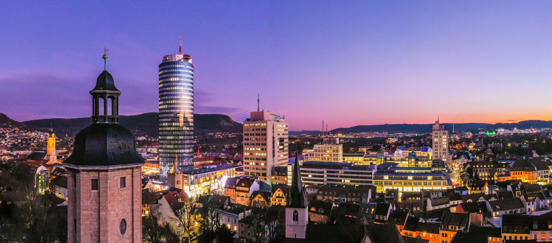 Abendliche Stadtansicht von Jena mit beleuchteten Gebäuden, dem markanten JenTower und einem Kirchturm im Vordergrund