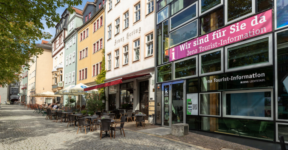 Straßenszene mit Außenbestuhlung vor historischen Gebäuden und moderner Glasfassade der Jena Tourist-Information mit sichtbarem Schriftzug 'Wir sind für Sie da Jena Tourist-Information'
