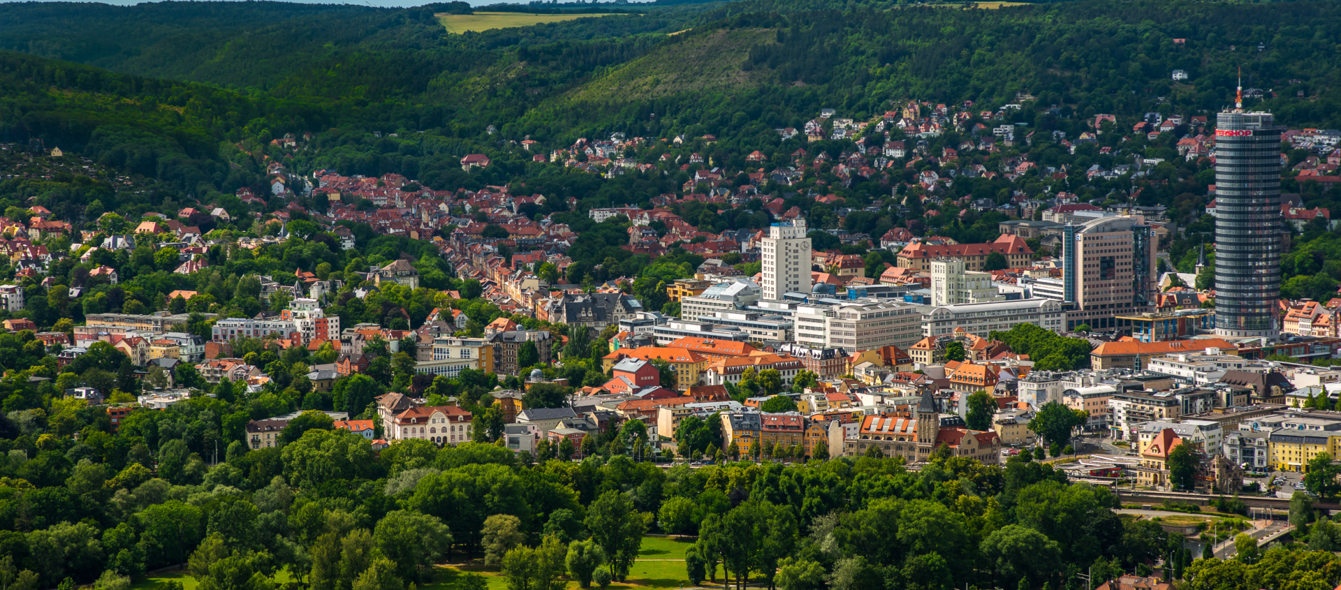 Panorama von Jena mit grünen Wäldern und Hügeln im Hintergrund, Stadtzentrum mit markantem JenTower und roten Dächern