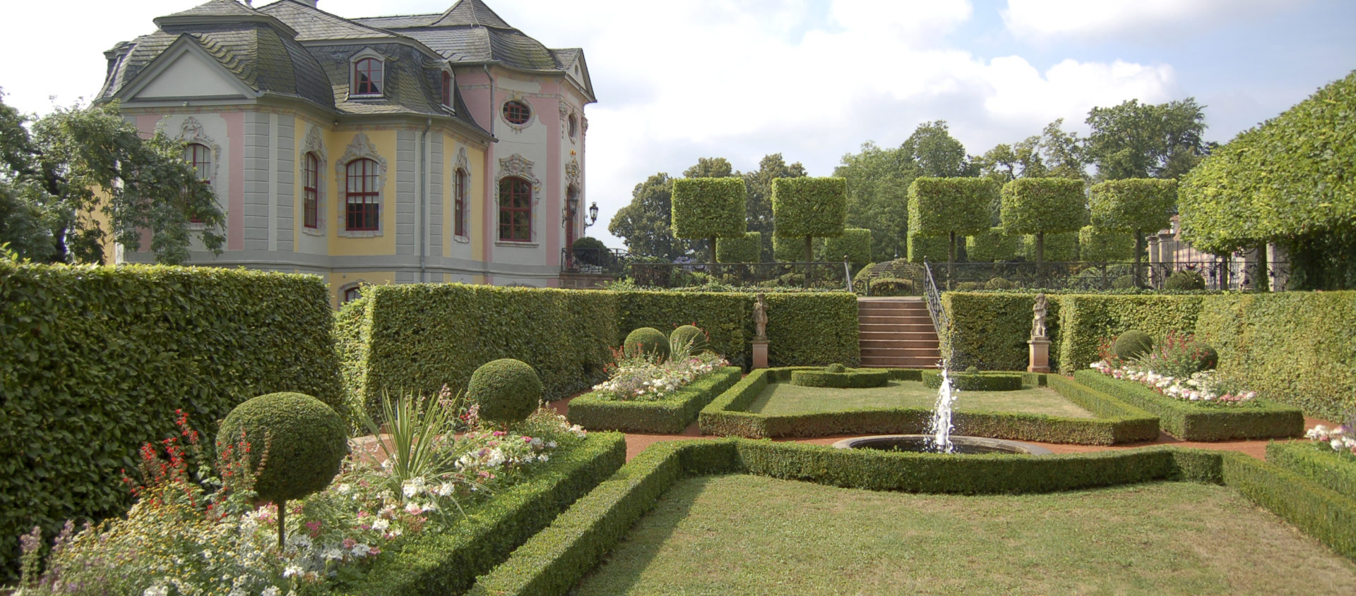 Barockes Schlossgebäude der Dornburger Schlösser mit geschwungenem Dach und symmetrischem, formell geschnittenem Garten mit Springbrunnen im Vordergrund