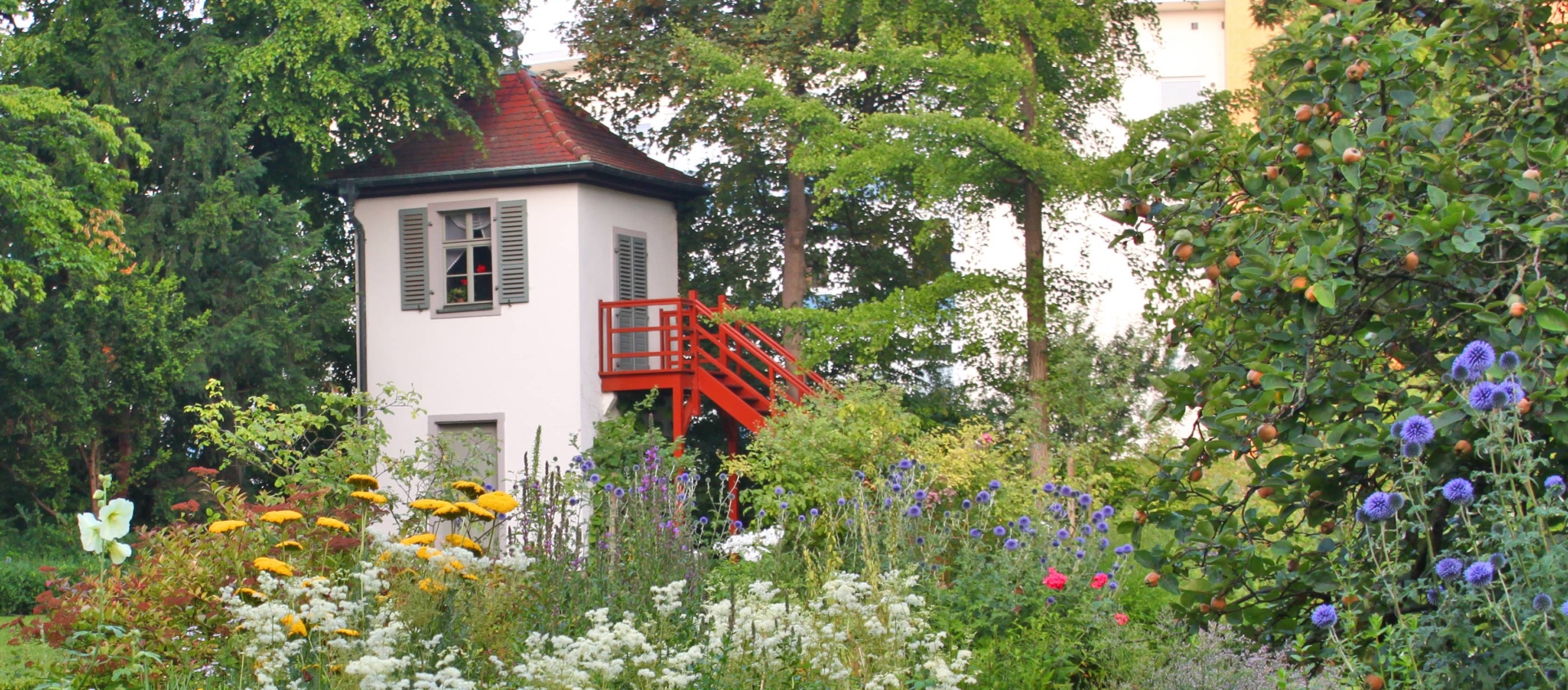 Kleines weißes Gartenhaus mit roter Außentreppe umgeben von vielfältiger, blühender Gartenpflanzen und Bäumen in Schillers Garten in Jena