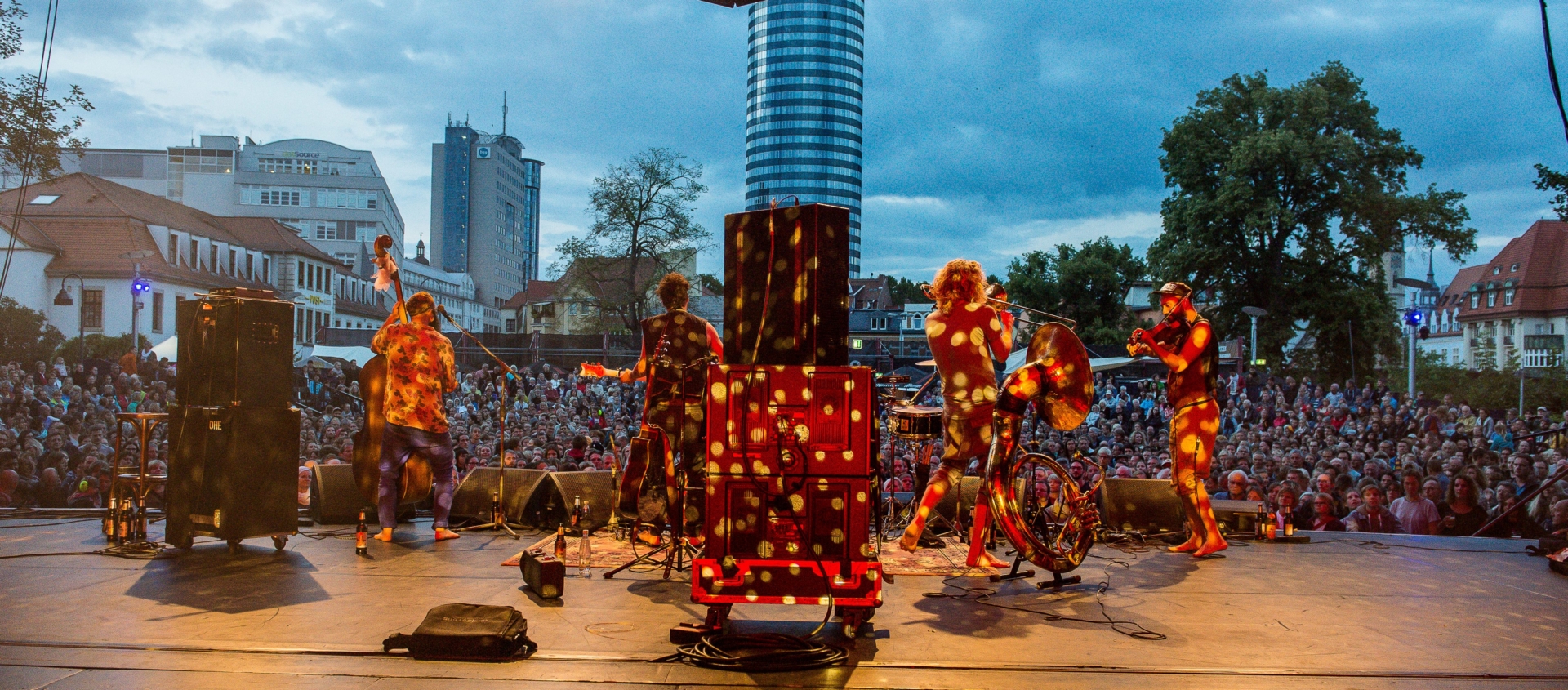 Blick von der Bühne auf eine Band mit mehreren Musikern und ein großes Publikum bei der Kulturarena Jena im Freien mit Stadtgebäuden im Hintergrund