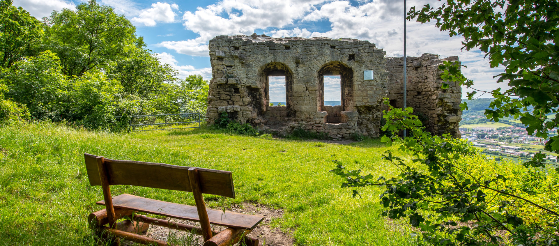 Ruine Kunitzburg mit drei Rundbogenfenstern auf einer Wiese mit einer Holzbank im Vordergrund und Blick auf die Stadt Jena im Hintergrund