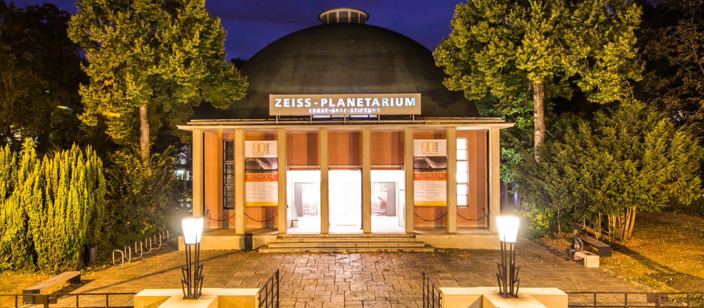 Das beleuchtete Zeiss-Planetarium Jena bei Nacht mit Kuppel, flankiert von Bäumen und einem gepflasterten Vorplatz mit Laternen