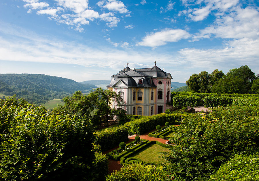 Barockes Schloss der Dornburger Schlösser umgeben von einem formalen Garten mit Hecken und Weinreben vor bewaldeten Hügeln unter blauem Himmel mit Wolken