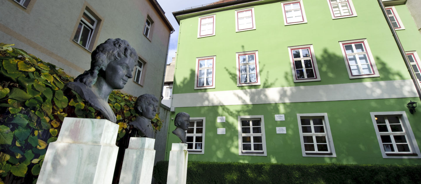 Drei Büsten auf weißen Sockeln stehen vor dem grünen mehrstöckigen Romantikerhaus in Jena