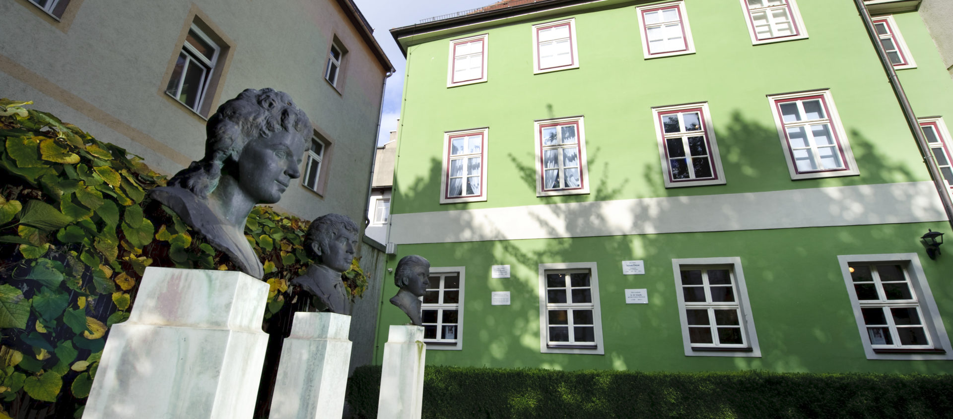 Drei Büsten auf weißen Sockeln stehen vor dem grünen mehrstöckigen Romantikerhaus in Jena