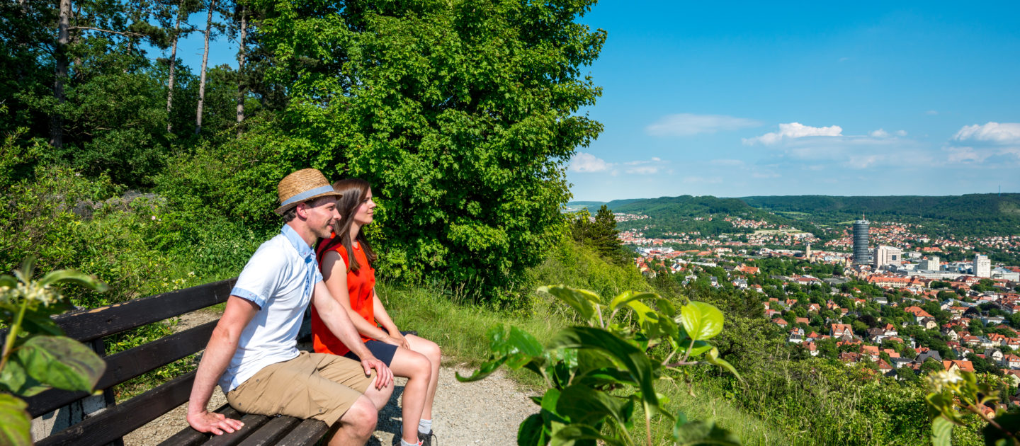 Zwei Personen sitzen auf einer Bank am Wanderweg Saalehorizontale mit Blick auf Jena und bewaldete Hügel unter blauem Himmel