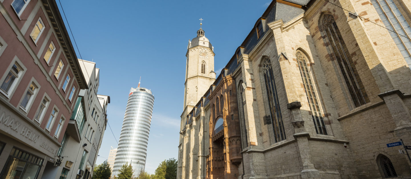 Blick entlang einer gepflasterten Straße auf die Stadtkirche St. Michael in Jena mit ihrem hohen Turm rechts und dem Jentower im Hintergrund links