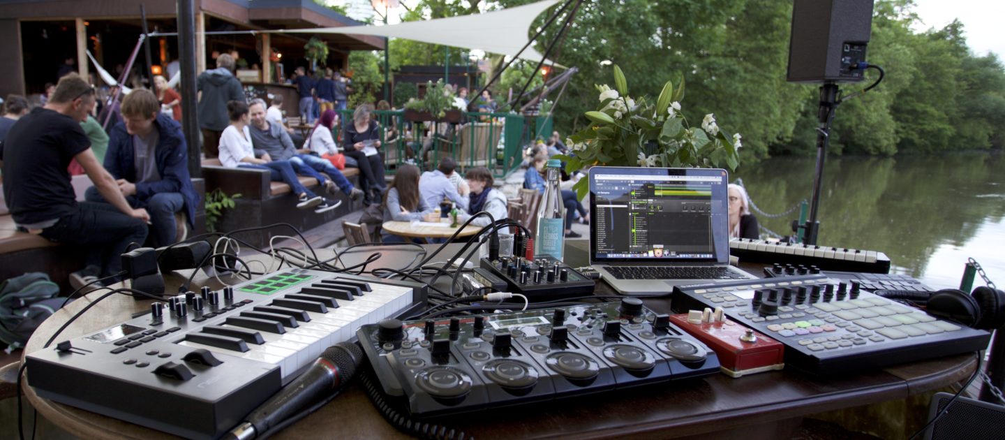 Musik-Equipment mit Mischpult, Laptop und Keyboard auf einem Tisch am Ufer der Saale, im Hintergrund sitzende Menschen und ein Pavillon am Strand 22 in Jena